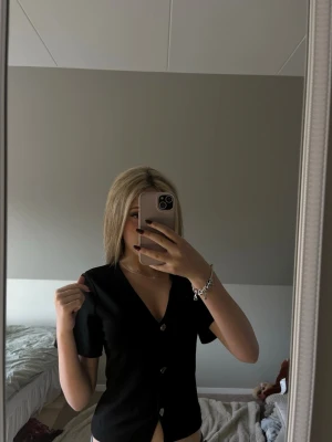 Svart v-ringad topp med knappar - Säljer en svart v-ringad topp med korta ärmar och knappar framtill. Enkel och stilren design som passar till många olika outfits. Perfekt för dig som gillar en clean look.