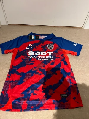 JDT matchtröja Nike röd/blå M - Snygg JDT matchtröja från Nike i rött och blått med kamouflagemönster och klubbmärke på bröstet. Tröjan har korta ärmar, krage och tryckta sponsorloggor framtill. Tillverkad i lätt och ventilerande polyester, perfekt för fotboll eller träning.