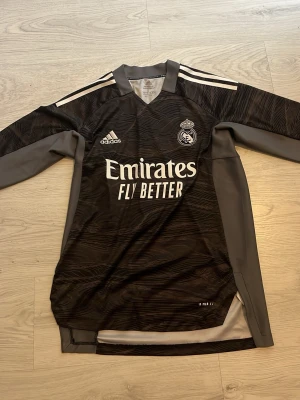 Real Madrid långärmad målvaktströja - Snygg svart och mörkgrå långärmad målvaktströja från Adidas för Real Madrid. Tillverkad i Primeblue-material med Parley Ocean Plastic. Tröjan har vita detaljer, klubbmärke och Adidas-logga på bröstet samt 'Emirates Fly Better' tryckt framtill.