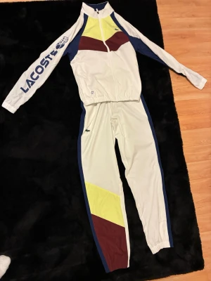 Lacoste vit träningsdress unisex - Snygg vit träningsdress från Lacoste med färgblock i gult, vinrött och blått. Jackan har dragkedja, hög krage och stor Lacoste-logga på ärmen. Byxorna har blå sidopaneler och klassisk krokodillogo. Perfekt för sport eller chill.
