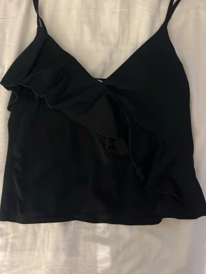 Svart volang topp med smala band - Säljer en svart linnetopp med smala axelband och volangdetalj framtill. Toppen är croppad och har en snygg v-ringning. Perfekt för dig som gillar en enkel men ändå trendig look.