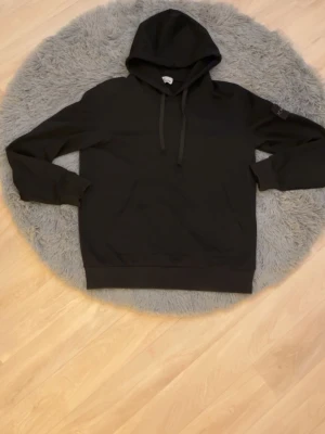 Svart hoodie från Stone Island - Säljer en svart hoodie från Stone Island med klassisk huva och dragsnören. Hoodien har en känguruficka framtill och ribbade muddar vid ärmslut och nederkant. Perfekt för dig som gillar stilrena och tidlösa plagg.
