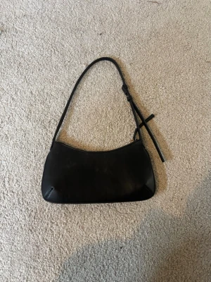 Svart axelremsväska i skinn - Snygg svart handväska i slätt skinn med justerbar axelrem och minimalistisk design. Väskan har en avlång, mjukt rundad form och stängs med dragkedja. Perfekt för dig som gillar stilrena accessoarer och vill ha en enkel men trendig look.