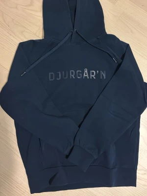 Mörkblå Djurgår'n hoodie - Mörkblå hoodie från Djurgårdens IF med trycket 'DJURGÅR'N' på bröstet. Klassisk huva med dragsko och magficka framtill. Officiell merchandise, perfekt för dig som vill representera DIF med stil. Priset går att diskutera!