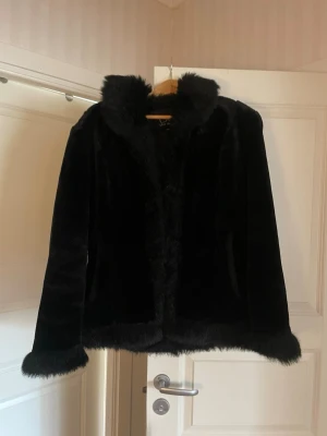 Svart fuskpälsjacka med krage - Snygg svart fuskpälsjacka med fluffig krage och detaljer vid ärmslut och nederkant. Jackan har rak passform och stängs framtill. Perfekt för att hålla värmen och ge en cool look under vintern eller hösten.