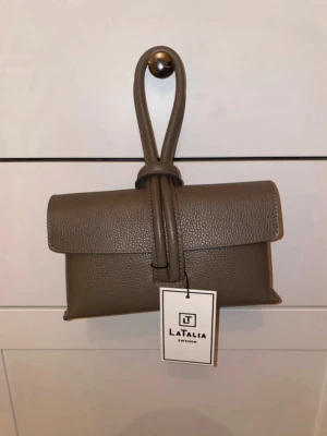 Brun axelväska från LataliA - Snygg brun axelväska från LataliA i stilren design. Väskan är tillverkad i strukturerat konstläder och har ett unikt knytband framtill. Justerbar axelrem för perfekt passform. Perfekt accessoar för dig som gillar minimalistisk och trendig stil.