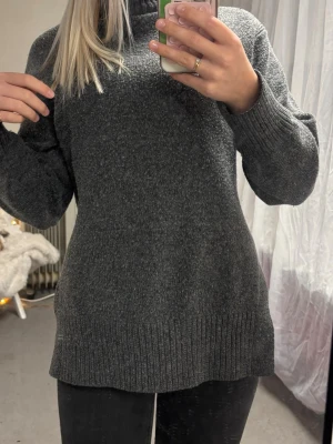 Mörkgrå stickad polotröja - Otroligt mysig mörkgrå stickad polotröja med lång ärm och ribbade muddar vid ärmslut och nederkant. Tröjan har en rak passform, slits på sidan och hög hals. 