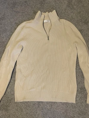 Beige stickad tröja Selected Homme - Snygg beige stickad tröja från Selected Homme med halv dragkedja och hög krage. Tröjan har lång ärm, ribbade muddar och är tillverkad i bomull. Perfekt för lager på lager och enkel att matcha med olika outfits.