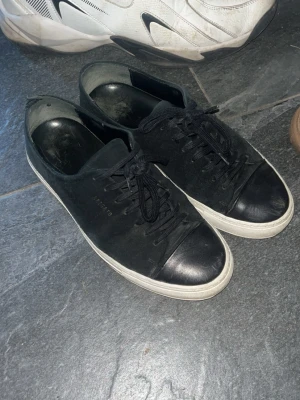 Axel arigato skor - Säljer ett par svarta sneakers från Axel arigato med klassisk design. Skorna har svart mocka på ovansidan, svarta snören och en vit sula. Tåpartiet är förstärkt med svart gummi för extra hållbarhet. Perfekta för dig som gillar stilrena och tidlösa sneakers. Används mycket och är helt ok i skicket. Dock ett jätte litet hål finns vid hälen inne i skon men inget som syns eller känns ❣️