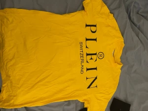 Gul t-shirt från Plein Switzerland - Snygg gul t-shirt från Plein Switzerland med stor svart logga framtill. Klassisk rund halsringning och korta ärmar. Tillverkad i mjuk bomull som känns skön mot huden. Perfekt för dig som vill sticka ut med färg.