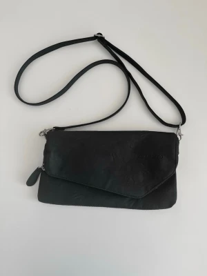 Svart multifunktionell väska från Björn Borg - Snygg svart axelremsväska / handväska / väska från Björn Borg med justerbar rem. Praktisk och smart modell som kan fällas upp för att bli större väska och fällas med för att bli mindre väska. Väskan har ett huvudfack som stängs med dragkedja och ett mindre fack med dragkedja på baksidan. Locket stängs med magnetknapp. Lite missfärgat tyg inuti, se bild, men annars i mycket gott skick.   Skickas mot porto, kan även hämtas i Stockholm. 