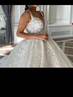 Vit lång spetsklänning - Elegant långklänning i vit spets med djup v-ringning och breda axelband. Klänningen har en voluminös kjol och vackra spetsdetaljer över hela plagget, vilket ger en romantisk och lyxig känsla. Perfekt för dig som vill sticka ut på bal eller fest.