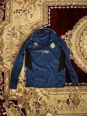 Blå träningsjacka Umbro Göteborg - Blå långärmad träningsjacka från Umbro med tryck från Elitfotbollsgymnasiet Göteborg. Jackan har dragkedja vid halsen, svarta paneler på sidorna och vita loggor. Materialet är troligtvis polyester, perfekt för träning eller uppvärmning.