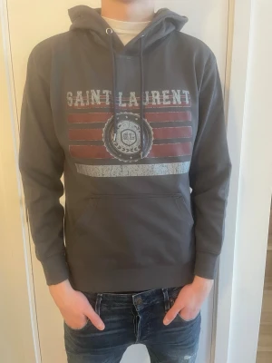 Grå Saint Laurent hoodie med tryck - Snygg grå hoodie från Saint Laurent med huva och dragsnören. Framsidan har ett vintageinspirerat tryck med röda och vita ränder samt ett SL-emblem i mitten. Klassisk känguruficka och ribbade muddar. Perfekt för en avslappnad streetwear-look.                 Bröstomfång: 98 cm Axelbredd: 44 cm Plagglängd: 63 cm Ärmlängd: 56 cm