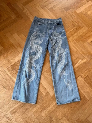 Blå jeans med drakmönster - Unika blå jeans med stora vita drakar tryckta längs båda benen. Byxorna har rak passform och hög midja, men ser också bra ut som low rise. Perfekta för dig som vill sticka ut med ett coolt motiv. Materialet är klassisk denim. Mått: midjemått 35 cm midjehöjden: 32 cm innebenslängd: 72 cm ytterbenslängd: 101 cm benöppning: 26 cm 