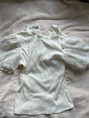 Vit topp med puffärmar och spets - Stl xs/s. Supersöt vit topp med ribbad struktur och puffiga spetsärmar med broderade blommor. Runt halsen finns en liten volangkant som ger en extra gullig touch. Perfekt för dig som gillar romantisk och trendig stil.