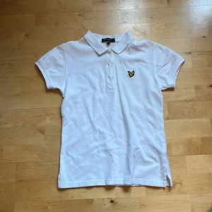 Vit pikétröja från Lyle & Scott M i barn - Klassisk vit pikétröja från Lyle & Scott med korta ärmar och gul broderad logga på bröstet. Tröjan har krage, knäppning med tre knappar och är tillverkad i mjuk bomull. Perfekt för en clean och sportig stil.