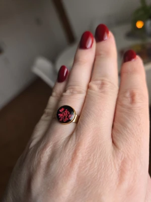 Ring med röda blommor - Unik guldfärgad ring med rund, svart platta dekorerad med röda blommor. Ringen har en enkel, stilren design och passar perfekt för dig som gillar detaljer med lite edge. Materialet är rostfritt stål.