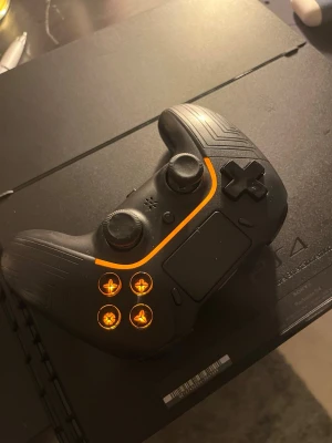 Svart trådlös handkontroll PS4-stil - Säljer en svart trådlös handkontroll med orange LED-belysning runt knapparna. Kontrollen har en modern design med texturerat grepp, två analoga spakar, styrkors, touchpad och klassiska PlayStation-knappar. Passar perfekt för gaming på PS4.