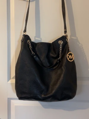 Svart Michael Kors handväska i läder - Säljer en svart handväska från Michael Kors i mjukt, kornigt läder. Väskan har guldfärgade kedjedetaljer och ett stort MK-hänge. Axelremmen är justerbar och väskan har en rymlig insida. Perfekt för dig som gillar klassisk och stilren design.