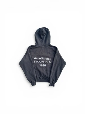 Svart hoodie från Acne Studios - Cool svart hoodie från Acne Studios med tryck på ryggen: 'Acne Studios STOCKHOLM 1996'. Klassisk design med huva och ribbade muddar. Perfekt för dig som gillar streetwear och minimalistisk stil.