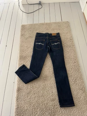 Mörkblå jeans från Nudie Jeans - Säljer ett par mörkblå jeans från Nudie Jeans med klassisk femficksmodell och kontrastsömmar. Jeansen har raka ben och tydliga Nudie-broderier på bakfickorna. Perfekta för en clean och stilren look. De är helt nya inte alls använda.