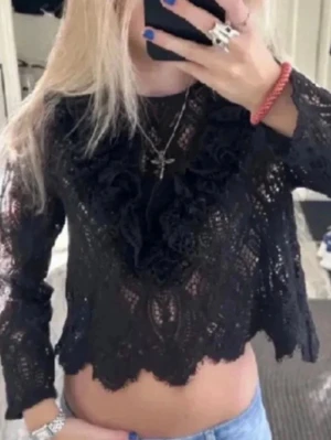 Svart spetsblus med volang - Säljer en svart, croppad spetsblus med långa ärmar och volangdetaljer framtill. Blusen har ett genombrutet mönster och en snygg scallopad kant nedtill. Perfekt för dig som gillar detaljer och vill sticka ut lite extra.