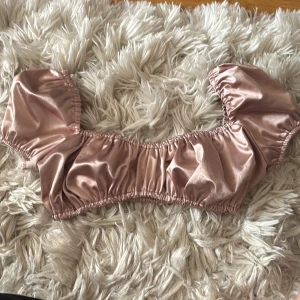 Champagnefärgad croppad blus i satin - Superfin croppad blus i glansig champagnefärgad satin med puffiga korta ärmar och resår både upptill och nedtill. Lite noprig på ena sidan (se bild) men går nog att få bort enkelt