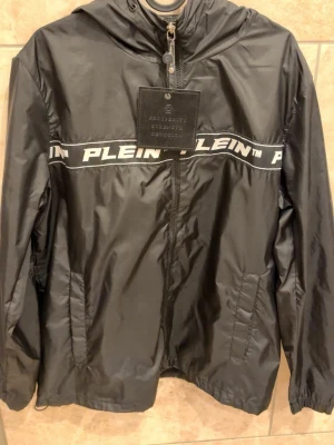 Svart vindjacka från Philipp Plein - Snygg svart vindjacka från Philipp Plein med huva och dragkedja framtill. Jackan har ett vitt logoband med texten 'PLEIN' över bröstet och två fickor framtill. Tillverkad i ett glansigt, lätt material som ger en sportig look. Ny pris är vid 6 tusen 