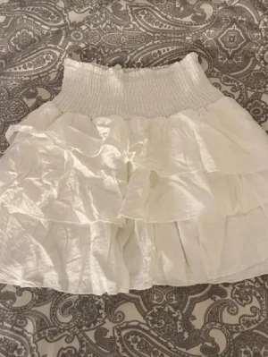 Volangkjol - Supersöt vit kortkjol med bred smock i midjan och flera lager volanger. Kjolen har en luftig och flowy känsla