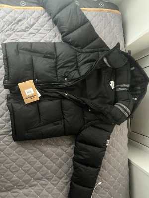 Svart pufferjacka från Burberry - Säljer en svart pufferjacka från Burberry med huva och dragkedja. Jackan har stora quiltade partier, sidofickor med lock och en klassisk Burberry-rutig insida på huvan. Perfekt för kalla vinterdagar och riktigt snygg streetstil.