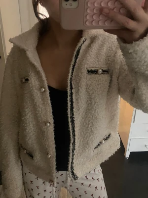 Vit teddyjacka med pärldetaljer - Supermjuk och fluffig vit teddyjacka med svarta detaljer och pärlknappar. Jackan har dekorativa fickor framtill med pärlor och svarta band, samt en klassisk krage. 