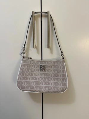 Beige axelväska från DKNY - Säljer en snygg beige axelväska från DKNY med logomönster över hela väskan. Väskan har vita detaljer, silverfärgad dragkedja och ett DKNY-emblem framtill. Axelremmen är vit och avtagbar. Perfekt storlek för det viktigaste och har ett innerfack med dragkedja.