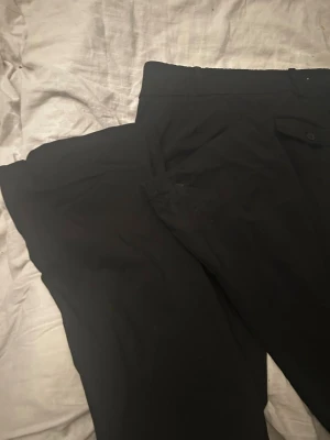 Svarta kostymbyxor från Bershka - Snygga svarta kostymbyxor från Bershka med raka ben och hög midja. Byxorna har två knappar i midjan och diskreta sidofickor. Perfekta för en clean och stilren look. Materialet känns mjukt och följsamt.