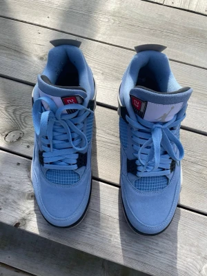 Air Jordan 4 University Blue Sneakers - Snygga Air Jordan 4 University Blue sneakers med ljusblå mocka, svarta och vita detaljer samt klassisk Jumpman-logga på hälen. Skorna har snörning och meshpaneler på sidorna för extra stil och komfort.