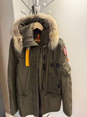 Olivgrön Parajumpers parkas med pälskrage - Snygg olivgrön parkas från Parajumpers med stor pälskantad huva och ikonisk gul rem vid halsen. Jackan har flera praktiska fickor, patch på ärmen och robust dragkedja. Perfekt för kalla dagar och riktigt street. Materialet är slitstarkt och vattenavvisande.