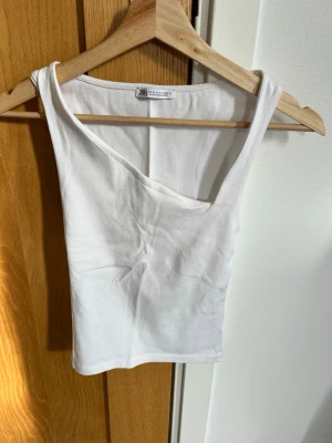 Vit asymmetrisk topp från Zara - Säljer en vit, croppad topp från Zara med cool asymmetrisk halsringning. Toppen är ärmlös och har en slim passform som framhäver siluetten. Perfekt för dig som gillar minimalistisk stil och vill ha något enkelt men ändå trendigt.