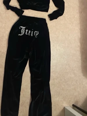 Svart velourset från Juicy Couture - Snyggt svart velourset från Juicy Couture med glittrande 'Juicy'-text i silver på både byxorna och ryggen av hoodien. Setet består av en croppad hoodie med långa ärmar och ett par mjuka byxor med resår i midjan. Perfekt för chill dagar hemma.