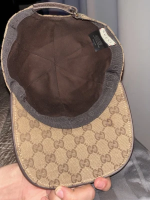 Beige keps från Gucci med GG-mönster - (pris går att diskuteras) Snygg beige keps från Gucci med klassiskt GG-monogram över hela kepsen. Justerbar rem baktill i brunt läder med präglad logga. Kepsen är tillverkad i canvasmaterial och har mörkbrun insida. Perfekt för dig som gillar exklusiva accessoarer (pris går att diskuteras)