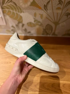 Vita Valentino sneakers med grön detalj - Säljer ett par vita Valentino sneakers med en bred grön rem över mitten. Skorna har lågt skaft, klassisk siluett och detaljerade nitar på hälen. Tillverkade i slätt läder för en clean och exklusiv look.
