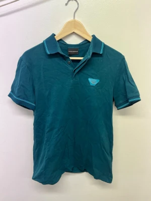 Blå pikétröja från Emporio Armani - Snygg blå pikétröja från Emporio Armani med klassisk krage och knappslå. Tröjan har korta ärmar och diskret logga på bröstet. Perfekt till jeans eller shorts för en avslappnad men stilren look. Den ändrar på framsidan från EA7 loggan till en smiley