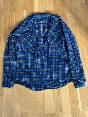 Blå rutig flanellskjorta (bluewear) - Snygg blå flanellskjorta med vitt och svart rutmönster. Skjortan har långa ärmar, två bröstfickor med knapp och klassisk krage. Perfekt för en avslappnad och trendig stil.
