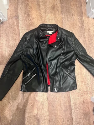 Svart skinnjacka med röda detaljer - Snygg svart skinnjacka från H&M med coola dragkedjor och quiltade axeldetaljer. Jackan har en röd innerfoder som ger en extra edge och ståkrage med tryckknapp. Perfekt för dig som vill ha en klassisk bikerstil.