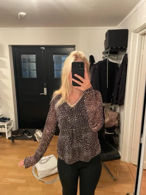 Leopardmönstrad blus med V-ringning - Snygg leopardmönstrad blus med V-ringning och glittrig beige kant runt halsen. Blusen har lång ärm och är tillverkad i ett tunt, lätt material som ger en avslappnad och trendig look. Perfekt för dig som vill sticka ut med ett djurmönster.
