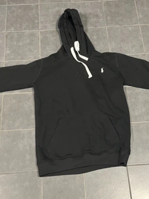 Svart hoodie från Polo Ralph Lauren - Snygg svart hoodie från Polo Ralph Lauren med vit logga på bröstet och vita dragsnören i huvan. Klassisk känguruficka framtill och ribbade muddar vid ärmslut och nederkant. Perfekt för en avslappnad och stilren look. Jag har knappt använt den på grund av att den är för stor för mig och storlek är S/M. Det är äkta polo kofta om nån undrar. Priset kan vi diskutera om