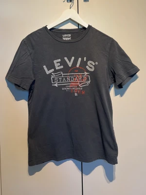 Mörkgrå Levi's t-shirt med tryck - Snygg mörkgrå t-shirt från Levi's med stort vitt och rött tryck på bröstet där det står 'Levi's Standard'. Klassisk passform och rund hals. Tillverkad i 100% bomull för en skön känsla. Perfekt till jeans eller shorts.