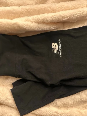 Svarta leggings från New Balance - Säljer ett par svarta leggings från New Balance med logga tryckt på låret. De har en tight passform och är tillverkade i ett stretchigt material som passar perfekt till träning eller chill. Klassisk design utan detaljer eller fickor.