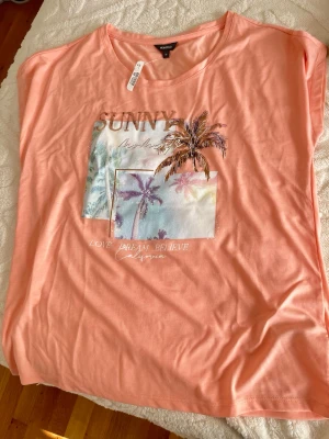 Persikofärgad t-shirt från Montego - Snygg persikofärgad t-shirt från Montego med tryck av palmer och texten 'SUNNY'. T-shirten har korta ärmar och en avslappnad passform. Perfekt för en somrig vibe och enkel att matcha med jeans eller shorts.