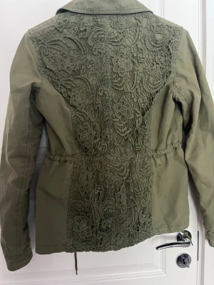 Olivgrön vindjacka med spetsdetaljer - Snygg olivgrön vindjacka från Hilfiger Denim med spetsdetaljer på ryggen och blommigt foder. Jackan har dragsko i midjan, två fickor framtill och tryckknappar vid kragen. Perfekt för dig som vill ha en unik och trendig jacka.