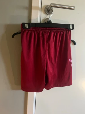 Röda sportshorts från Hummel - Snygga röda sportshorts från Hummel med elastisk midja och klassiska Hummel-pilar i ljusrosa på sidorna. Perfekta för träning eller sportiga dagar. Lätta och bekväma med en loose passform.